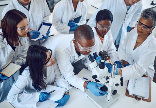 An overhead shot shows a diverse group of four female and two male scientists in lab coats and safety glasses surrounding and focusing on a black male colleague operating a compound microscope.
