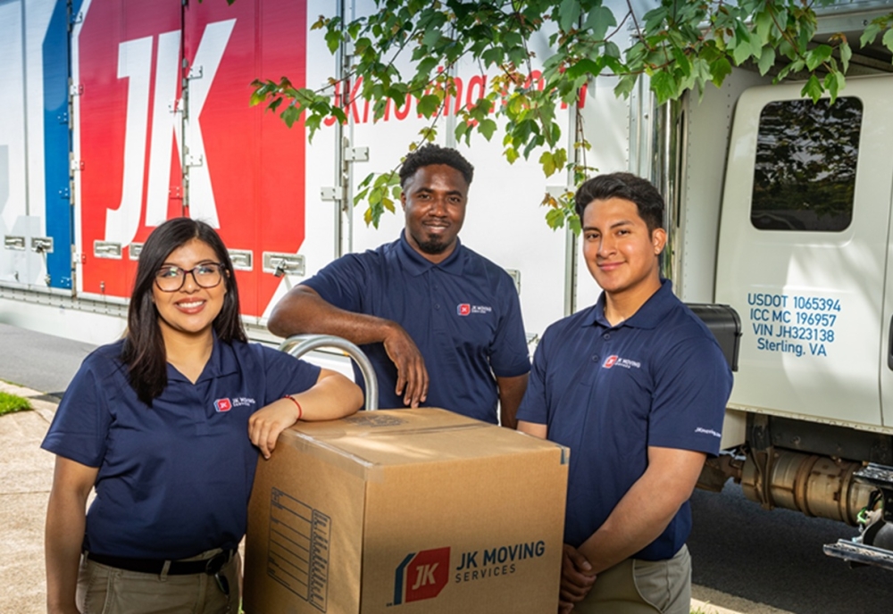 Three JK Moving Services team members wearing navy company polos stand beside a moving box in front of a JK moving truck, smiling outdoors before a residential move.