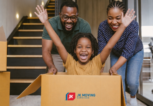 Excited child with arms raised sits in a JK Moving box while parents play in their new home