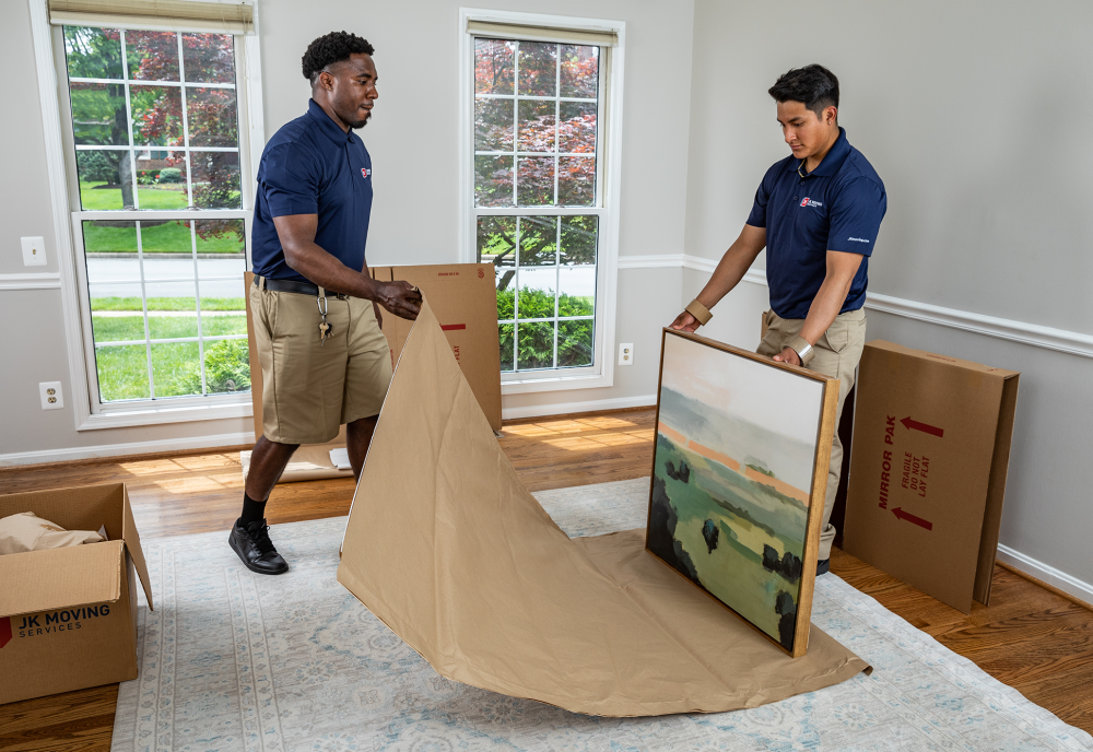 Two JK Moving crew members wrapping a framed landscape painting in protective paper inside a client’s home